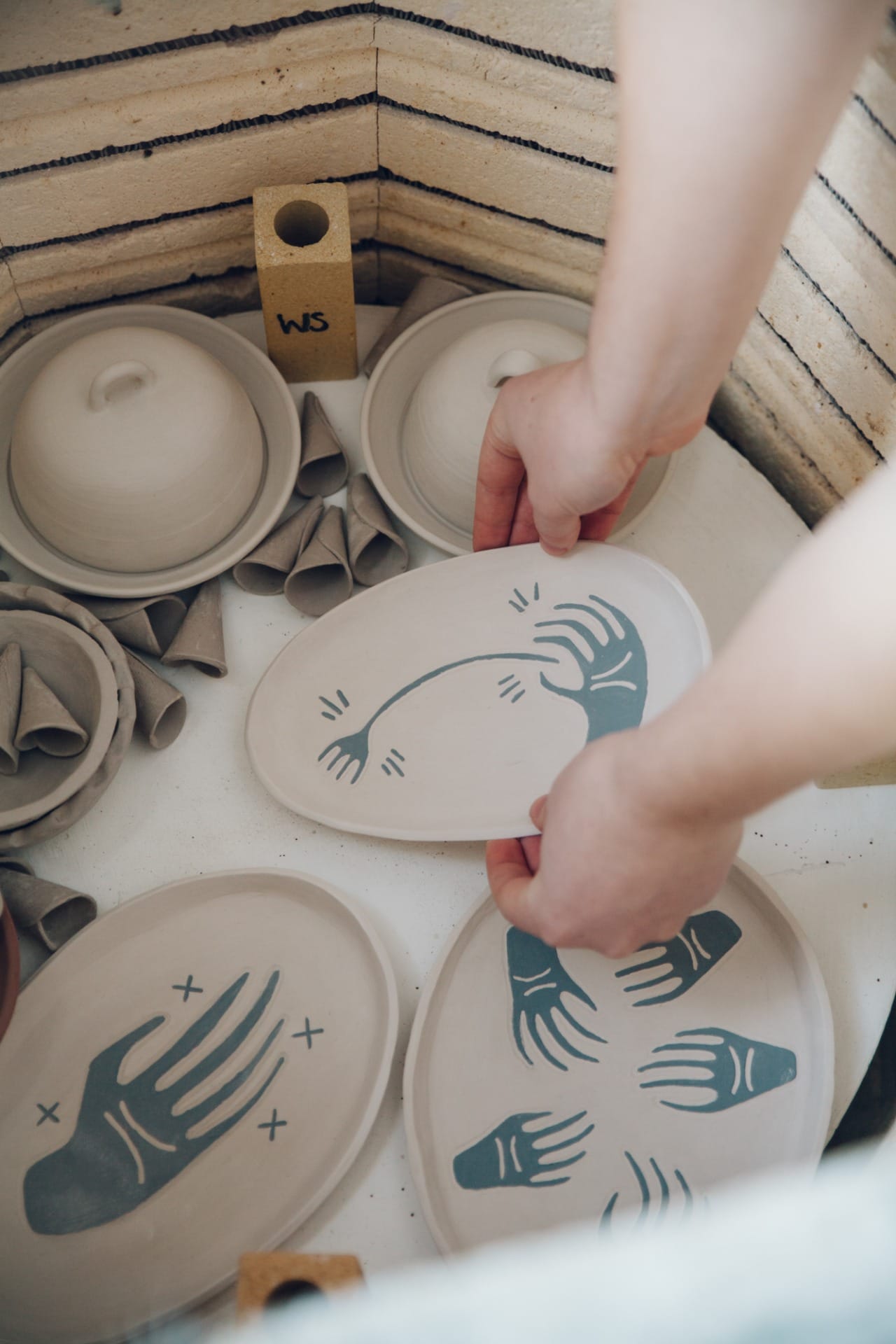 Hands loading handmade plates painted by Larissa James into an electric kiln.