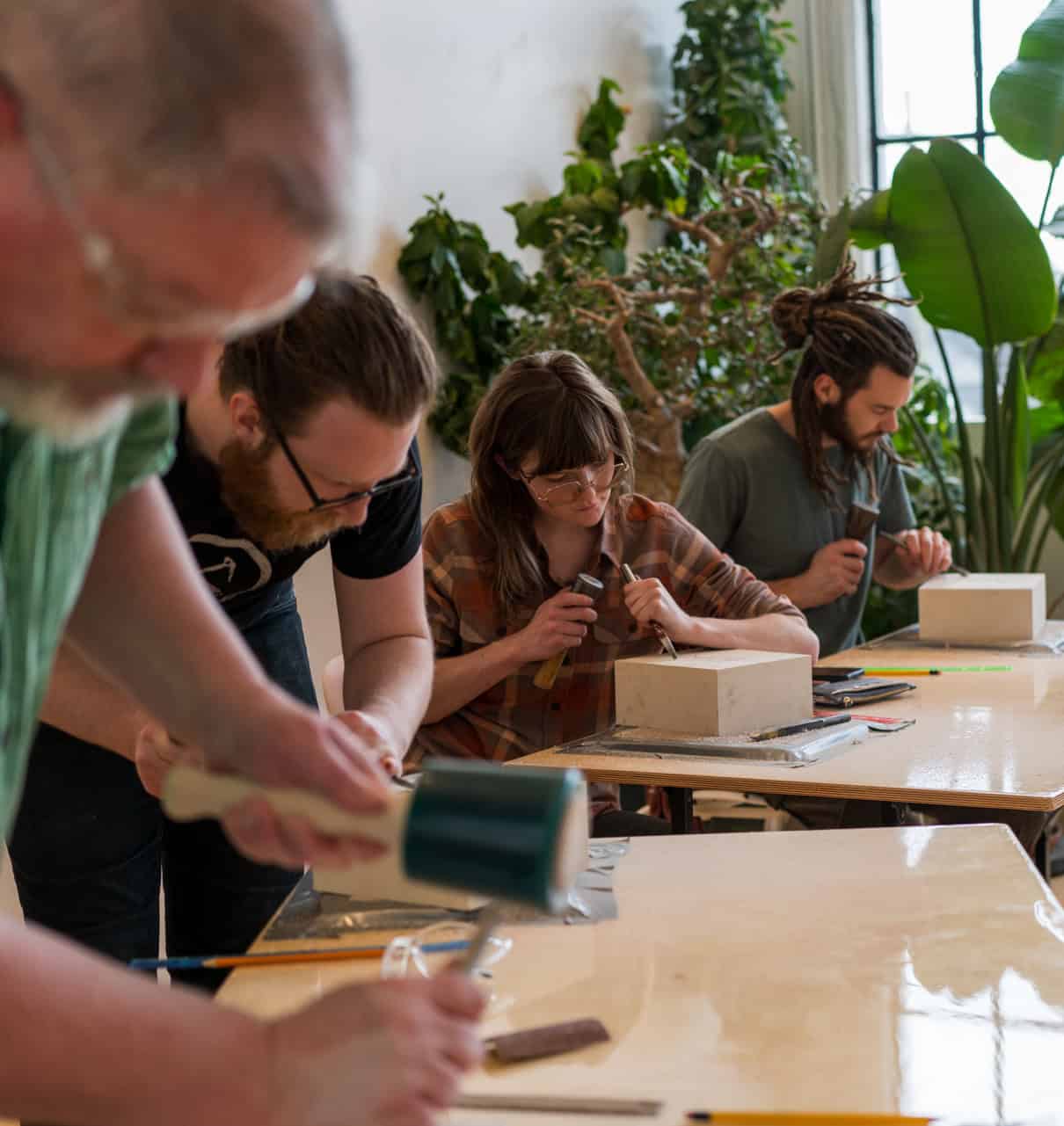 Four participants carving stone in a bright, plant-filled workshop, engaged in a stone carving class.